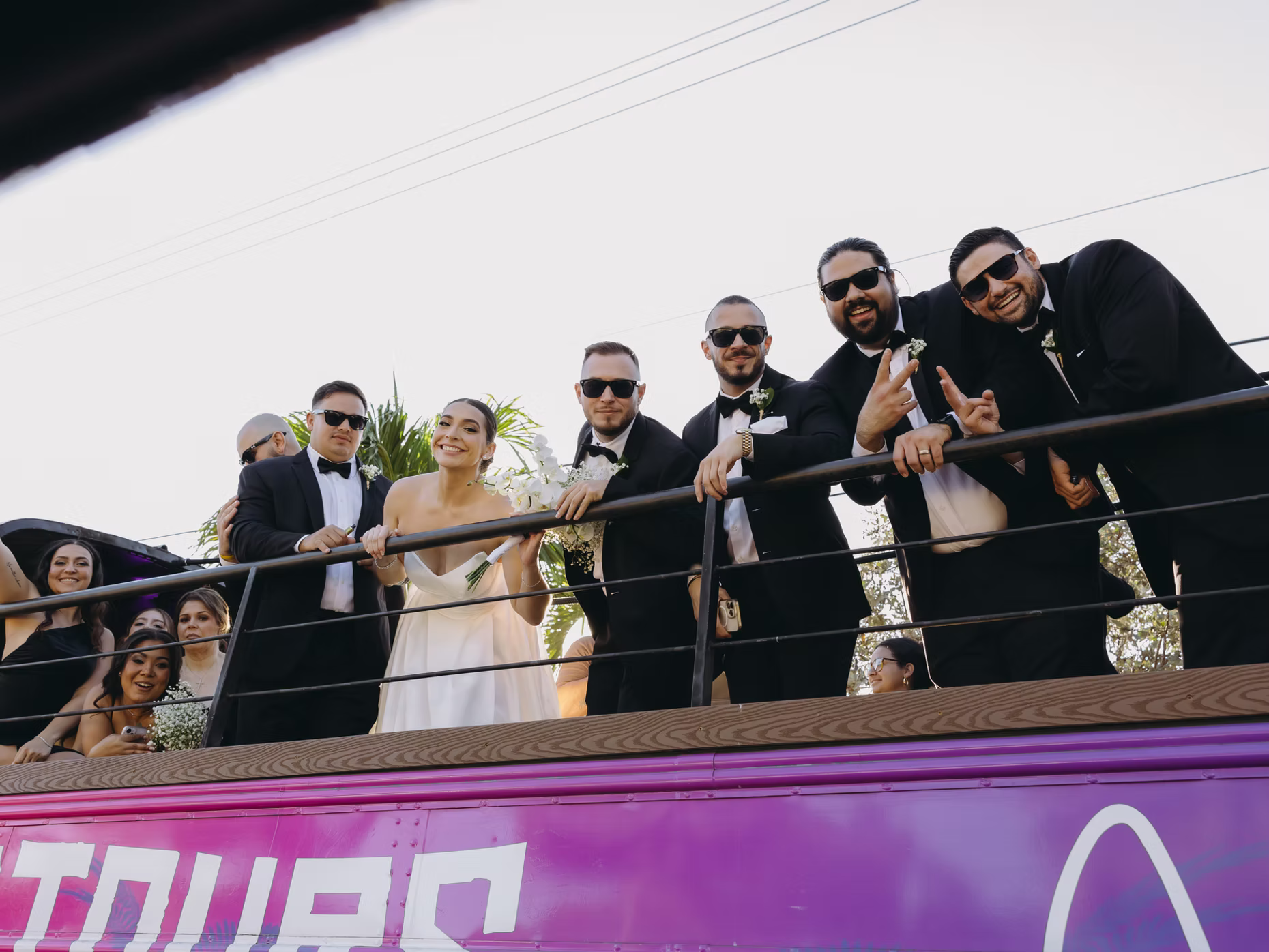 Bride and groom, along with their wedding party, standing on a pink RumbaTours open-air party bus, smiling and celebrating.